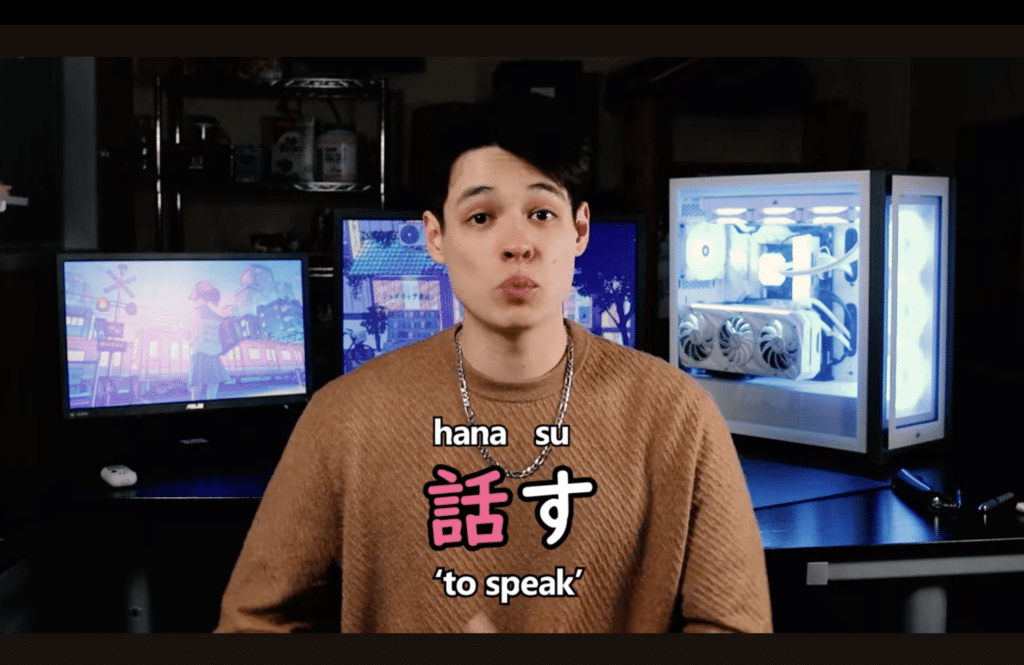 A man sitting with two computers behind him and Japanese letters o the screen